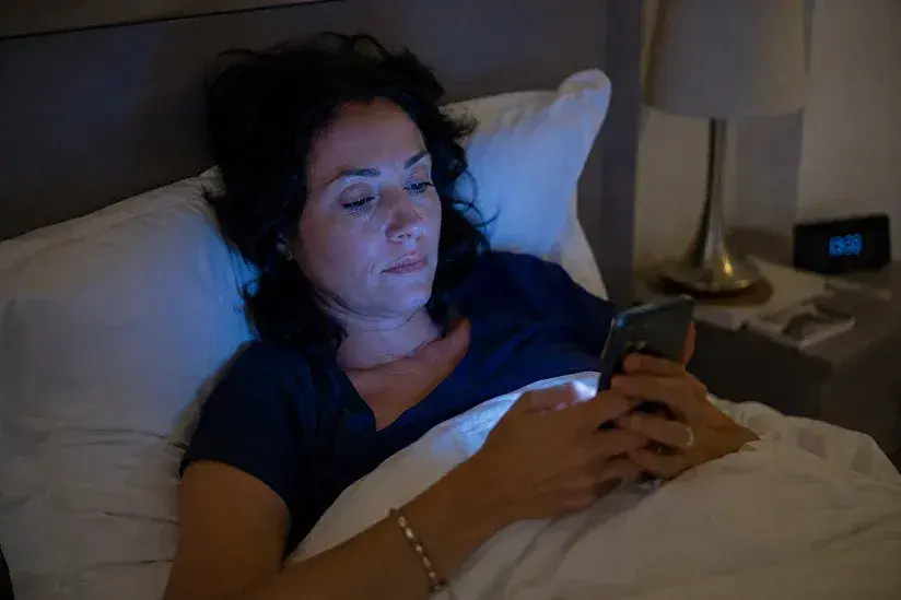 Une femme dans son lit, éclairée par la lumière bleue de son téléphone, perturbant son sommeil.