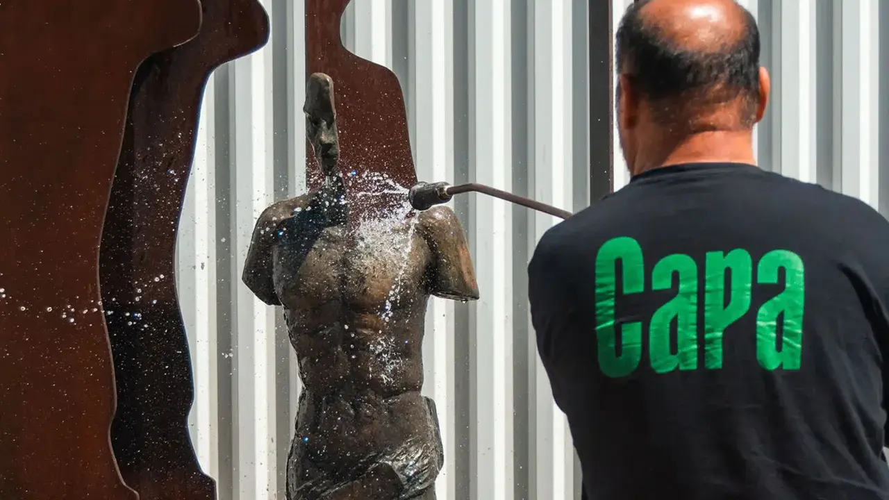 Hombre con camiseta negra con letras verdes "Capa" limpia una estatua de bronce con una manguera a presión.