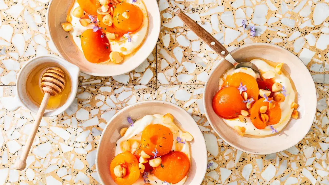 Trois bols de compote abricot onctueuse, garnie d'amandes et de fleurs violettes. Un pot de miel avec sa cuillère est à côté.