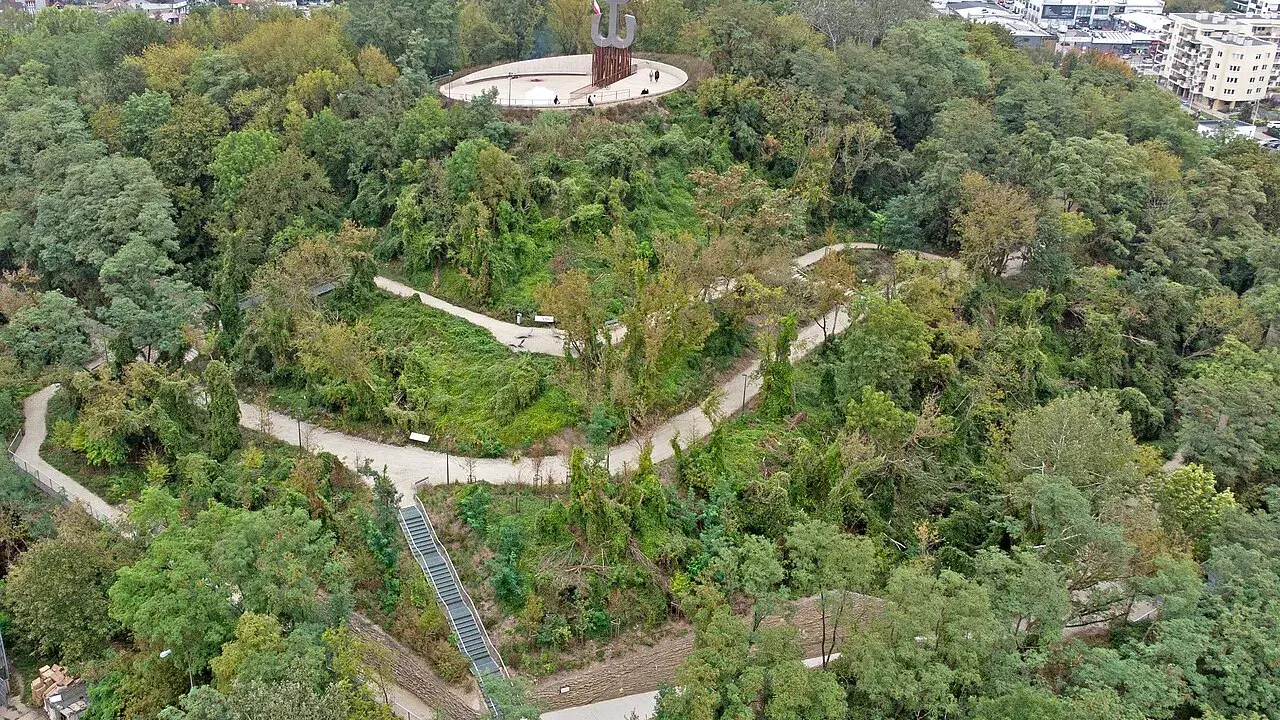 Wzgórze z pomnikiem Kotwicy, otoczone zielenią. Kręte ścieżki i schody prowadzą przez park akcji "Burza", oferując widok na miasto i Wisłę.