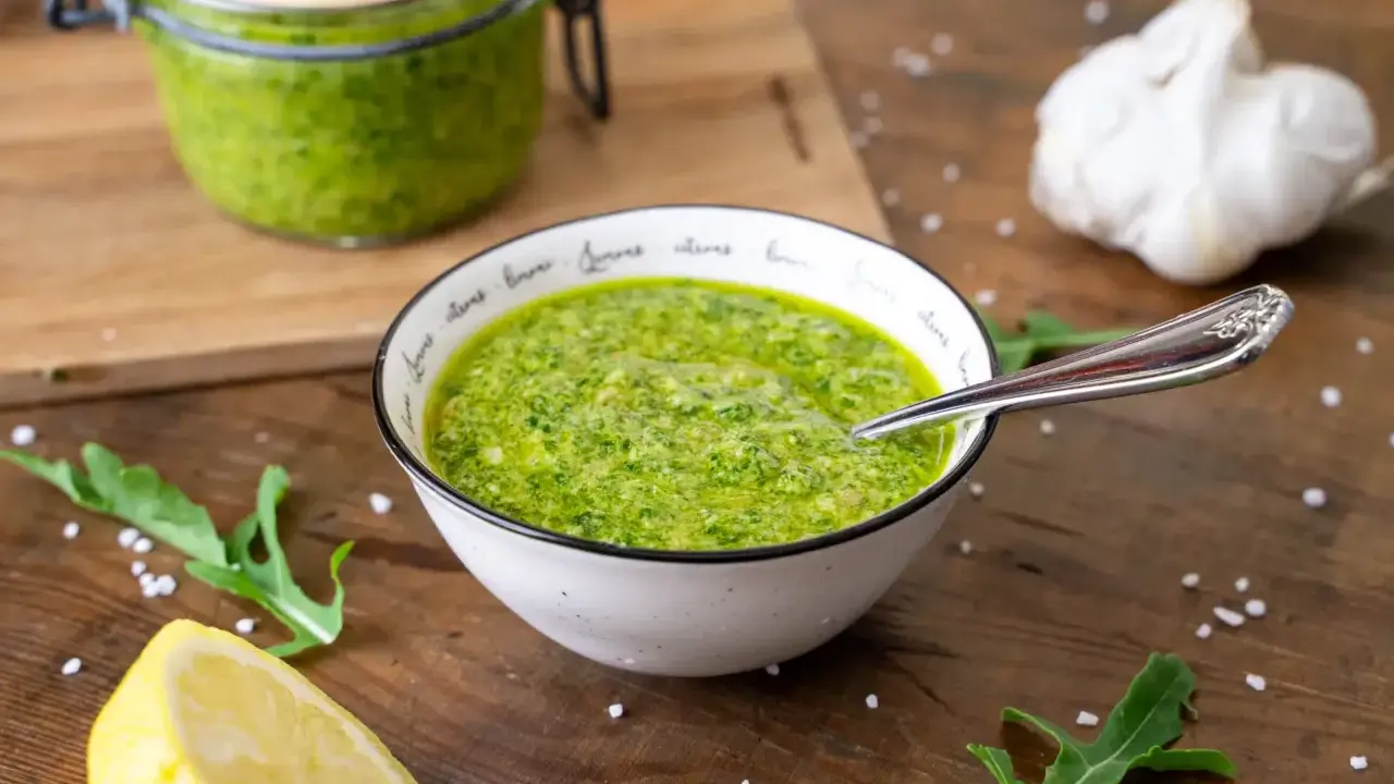 Frisches Rucola Pesto in einer Schüssel, daneben Zitrone, Knoblauch und Rucolablätter.
