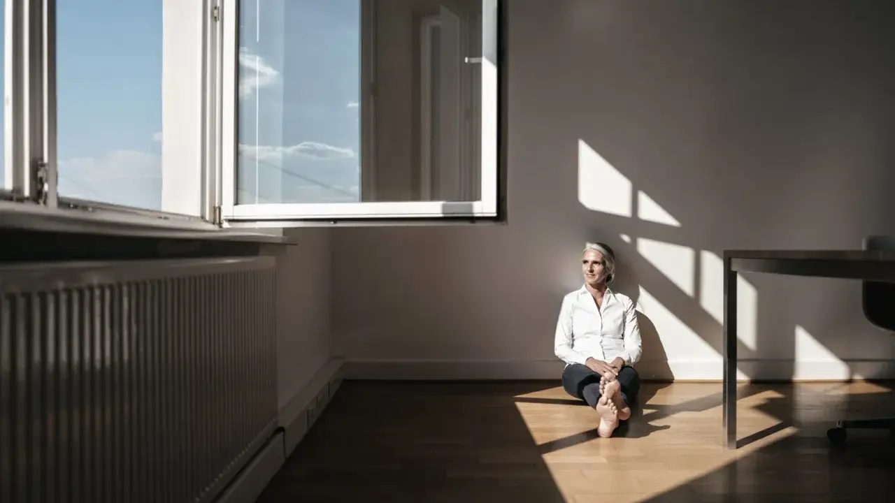 Kobieta w białej koszuli siedzi na podłodze w pustym pokoju, ciesząc się światłem wpadającym przez okno. To kwintesencja minimalizmu w domu.
