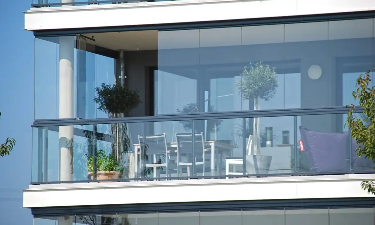 Un balcone moderno con vetrate scorrevoli, ideale per chiudere un balcone fai da te. Tavolo, sedie e un pouf viola creano un angolo relax.