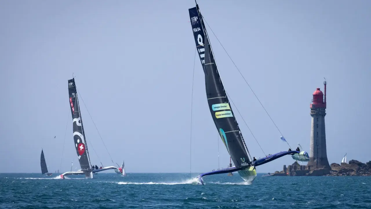Le trimaran Koesio fend les vagues, une course intense près d'un phare.