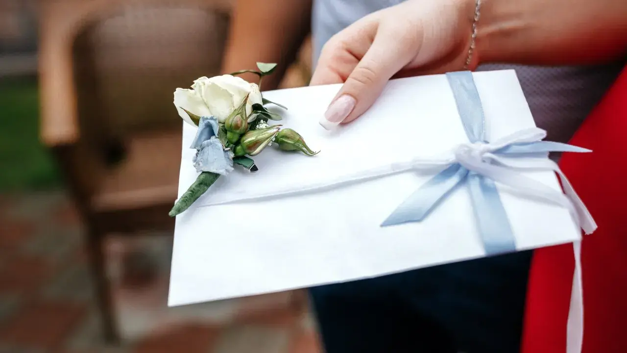 Biała koperta z błękitną wstążką i butonierką, symbolizująca pieniądze z wesela na konto.