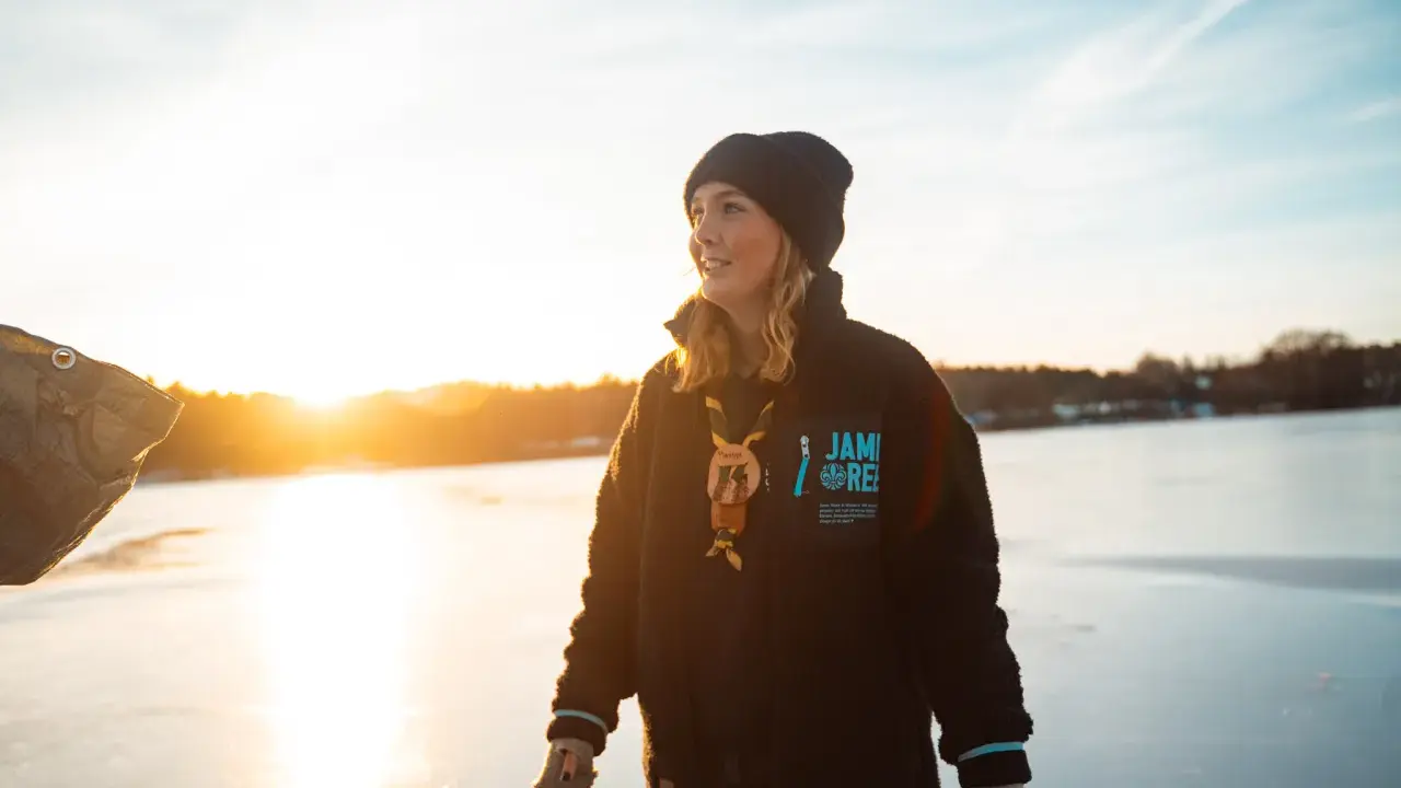 Young woman in a black beanie and fleece jacket standing on a frozen lake at sunset.