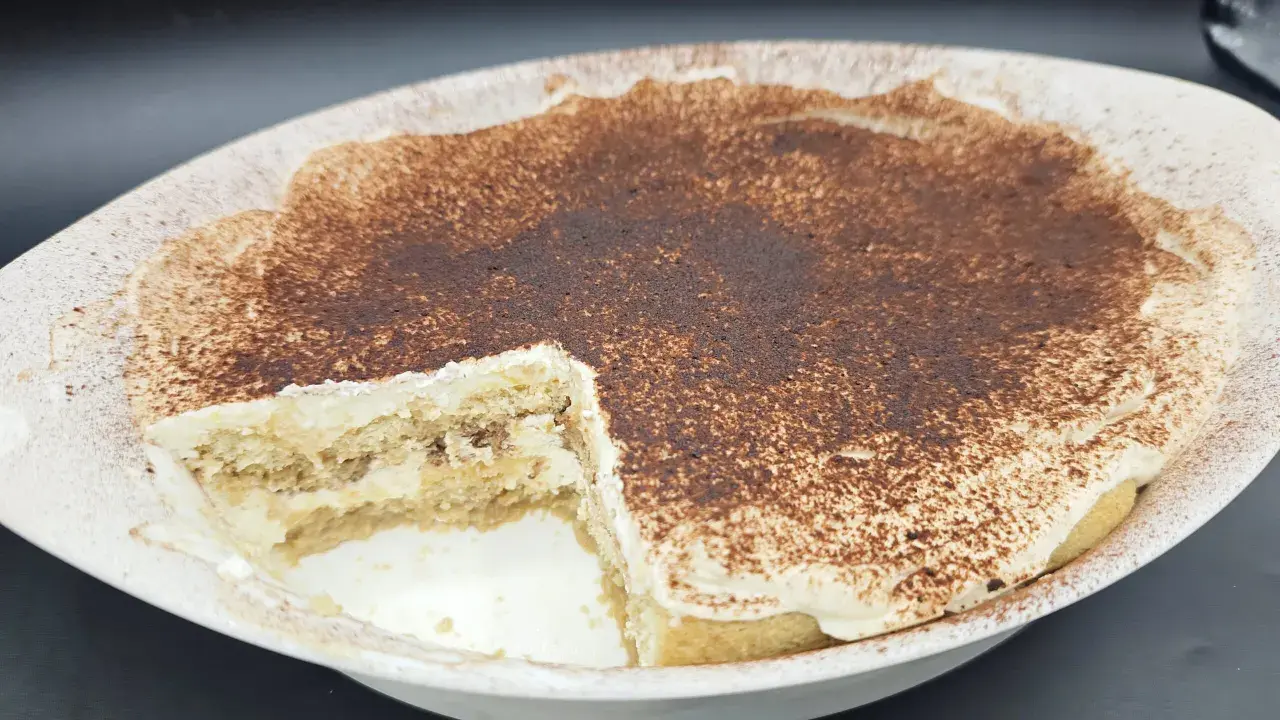 Un délicieux tiramisu sans oeuf, crémeux et saupoudré de cacao, dans un plat blanc. Une part a été retirée, révélant les couches de biscuits et de crème.