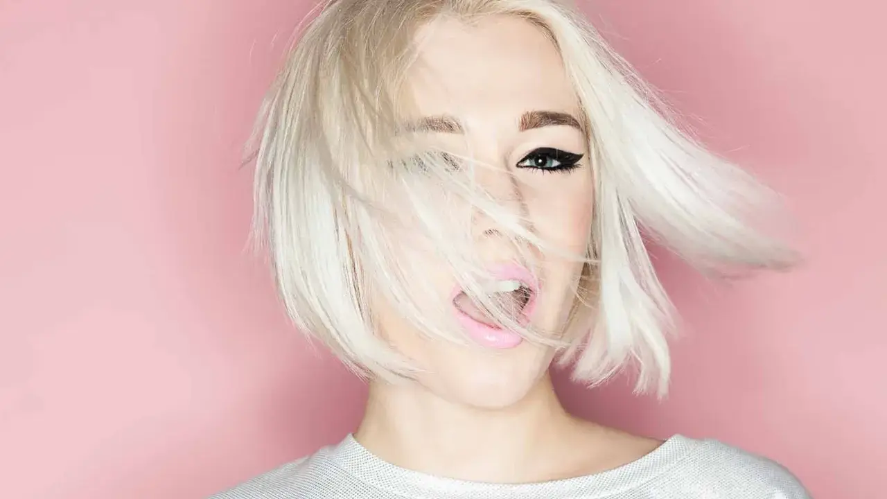 Mujer con cabello rubio platino y corte recto al hombro, con un ojo visible y labios rosados abiertos.