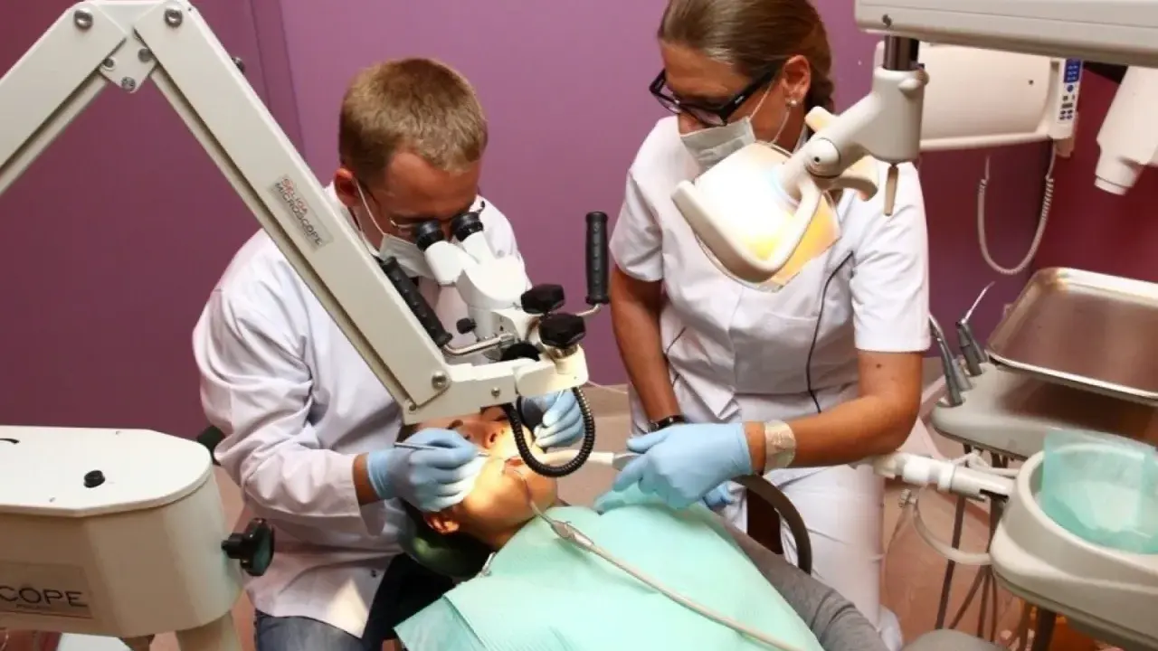 Dwaj dentyści w białych fartuchach pracują przy pacjencie, używając mikroskopu stomatologicznego.