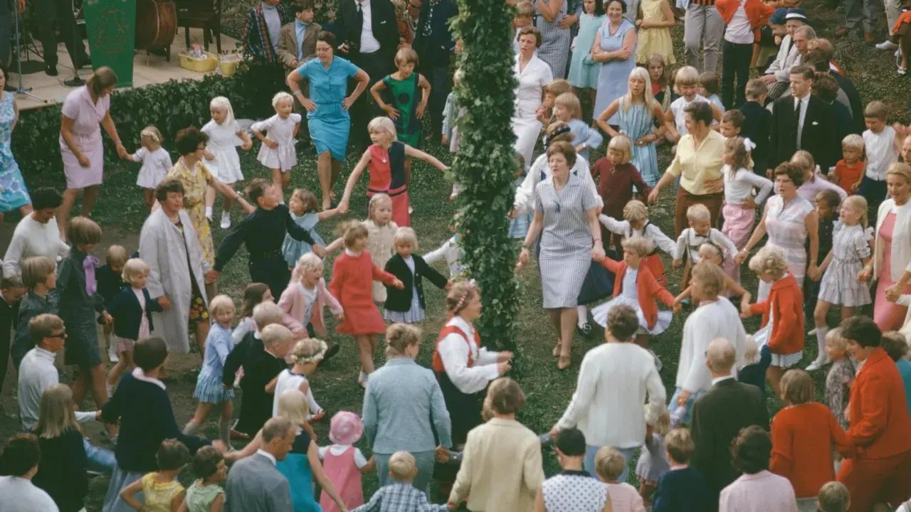 Många barn och vuxna dansar ringdans runt en midsommarstång i Sverige.