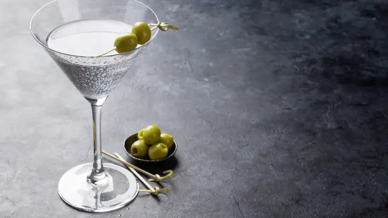 Ein Glas trockener Wermut mit Oliven garniert, daneben eine Schale mit weiteren Oliven auf dunklem Untergrund.