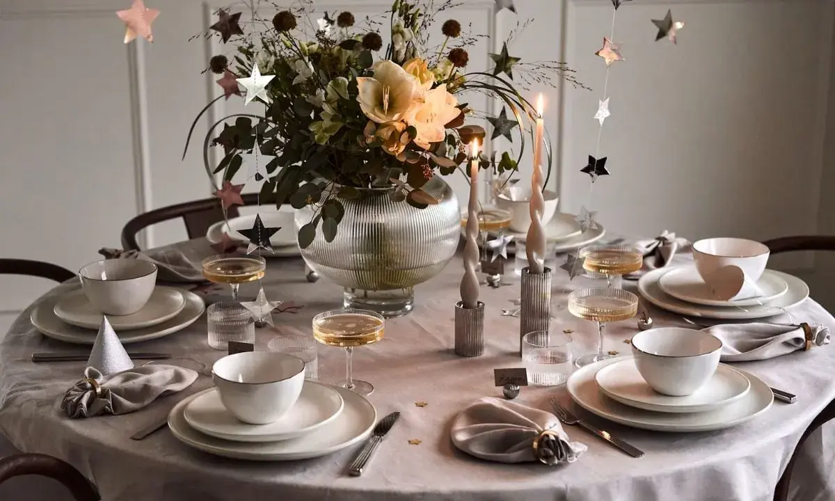 Enkel nyårsdukning med vita tallrikar, champagneglas, stjärnformade dekorationer och en blomsterbukett i en glaskaraff.