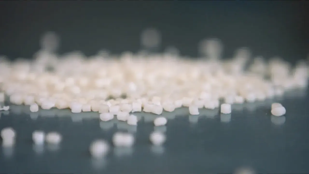 Små vita granulatkorn, råmaterial för nedbrytbara plastpåsar, ligger utspridda på en mörk yta.