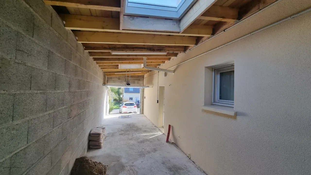 Vue d'une extension en cours de construction avec reprise de toiture, poutres apparentes et un puits de lumière.