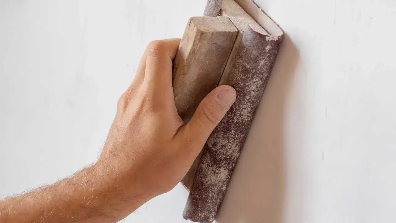 Mano lijando paredes para pintar. El bloque de madera con lija se desliza sobre la superficie blanca, preparándola para un acabado perfecto.