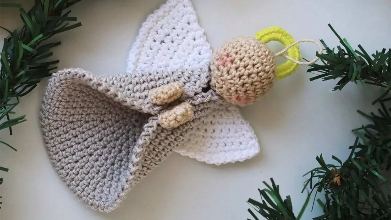 Un tierno angel amigurumi de ganchillo, con alas blancas y un cuerpo gris, adornado con ramitas verdes.