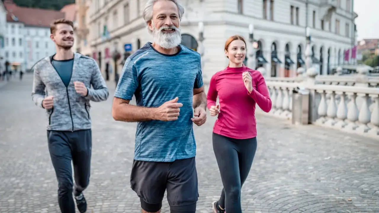 Un uomo con barba bianca e due giovani corrono sorridenti in città. Un esempio di come iniziare a correre a 50 anni sia possibile e gratificante.