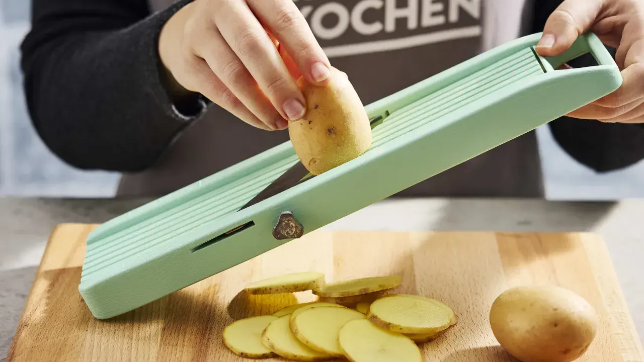 Eine Hand schneidet Kartoffeln für ein Kartoffelsandwich auf einem Hobel. Dünne Scheiben liegen auf dem Holzbrett.