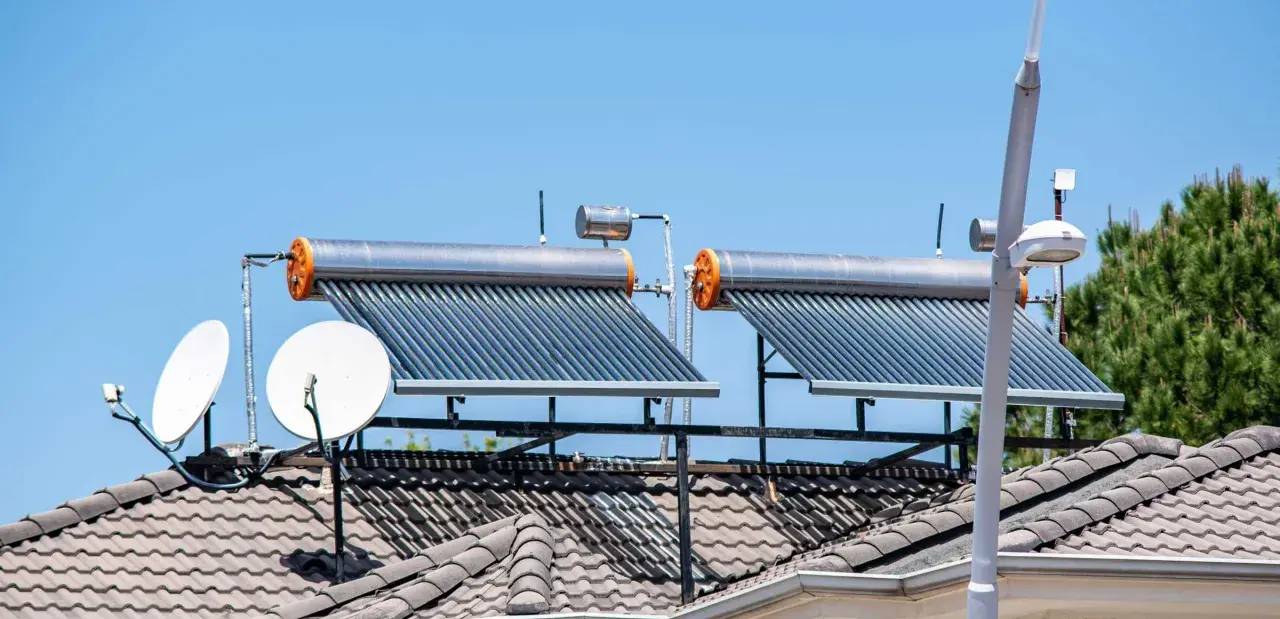Dos calentadores de agua caliente solar en un tejado, junto a antenas parabólicas, bajo un cielo azul.