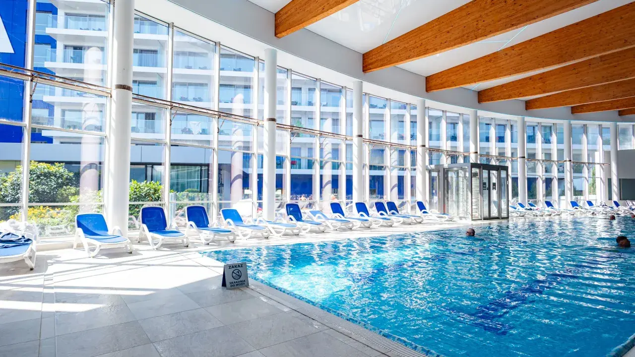 Wnętrze z basenami hotelowymi, leżakami i dużymi oknami z widokiem na budynek hotelowy.