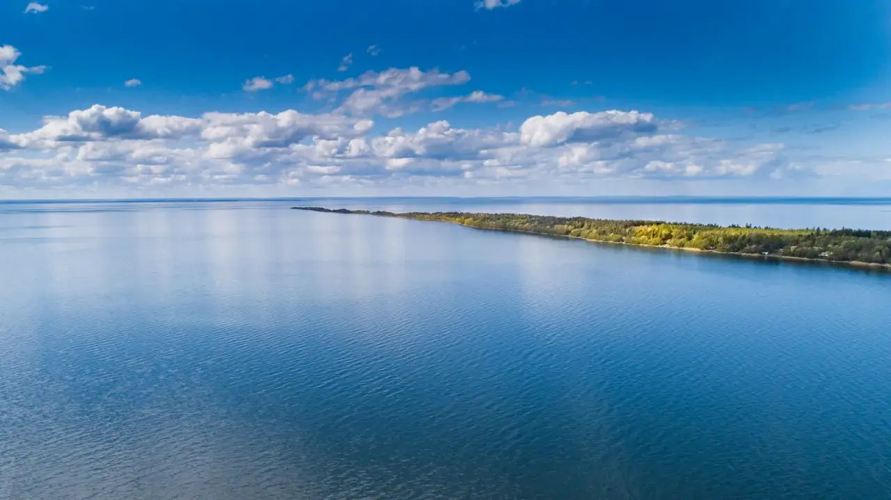 Blå himmel med moln över ett lugnt vatten. En lång, grönskande udde sträcker sig ut. Perfekt för vandring Mariestad.