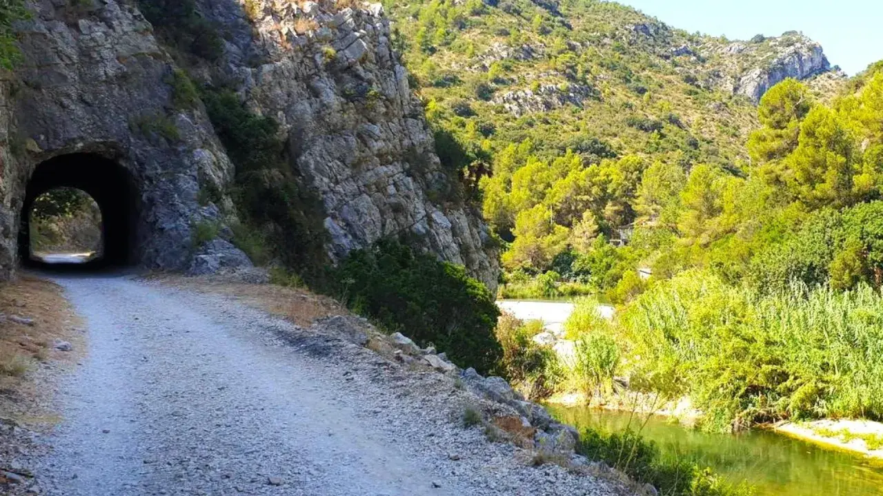 Camino de grava junto a un río y un túnel en la vía verde de Alicante. Naturaleza exuberante y rocas escarpadas.