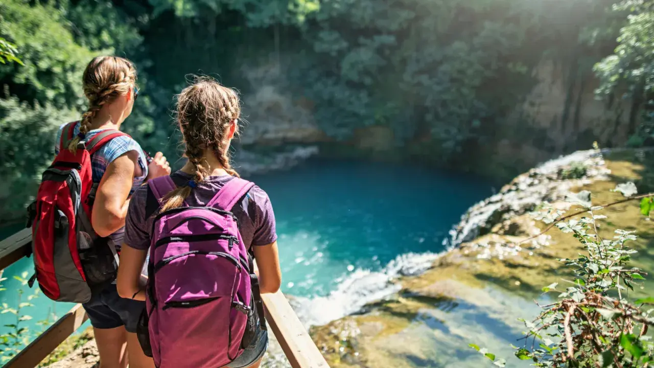 Due escursioniste con zaini ammirano una cascata turchese durante il loro trekking in Toscana.