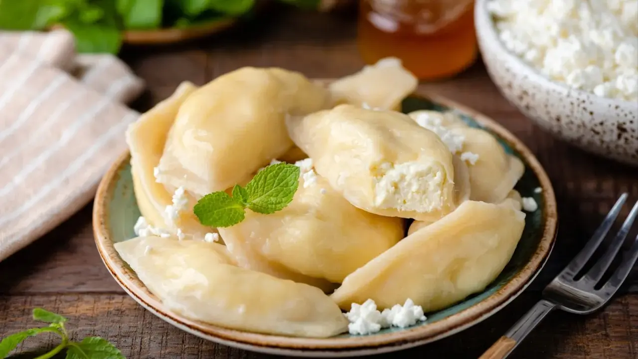 Pyszne pierogi z serem na słodko, posypane twarogiem i udekorowane miętą. Obok słoiczek miodu i miseczka z twarogiem.