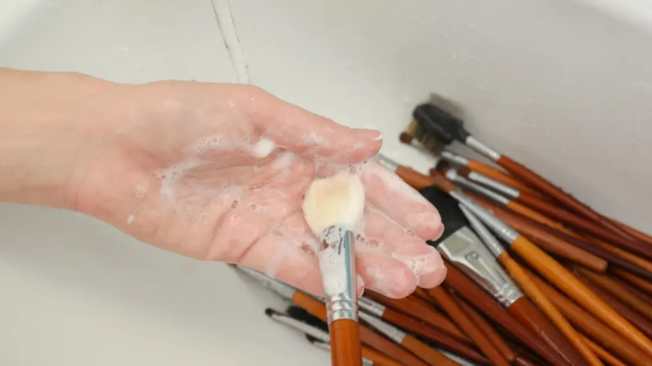 Mano con espuma jabonosa limpiando brochas de maquillaje en un lavabo.