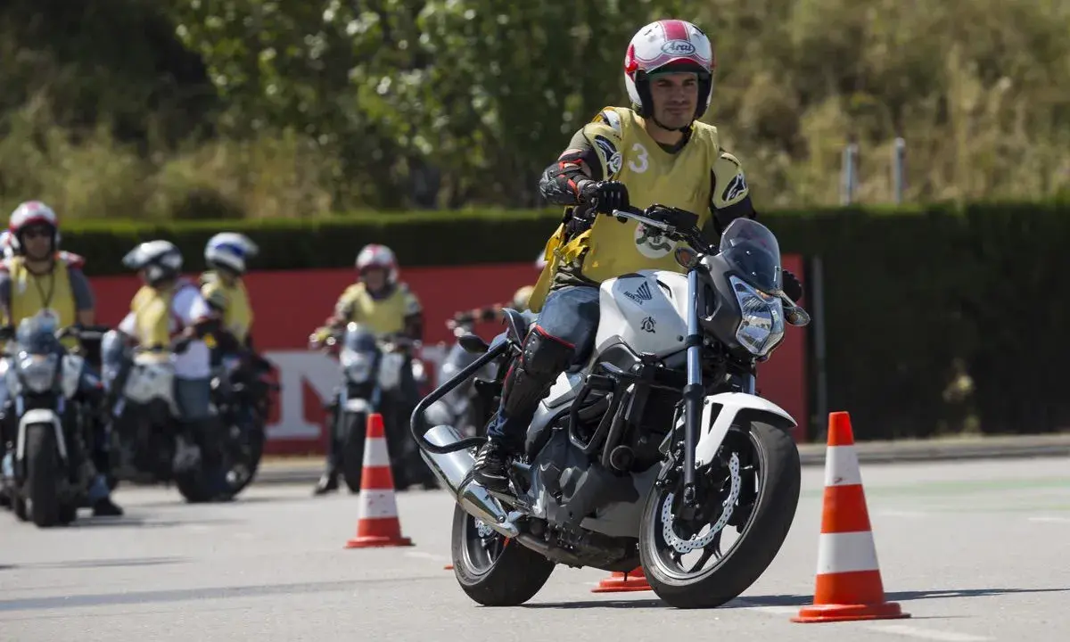 Hombre en moto blanca, con chaleco amarillo y casco, practicando maniobras en un curso carnet A. Otros motoristas y conos en el circuito.