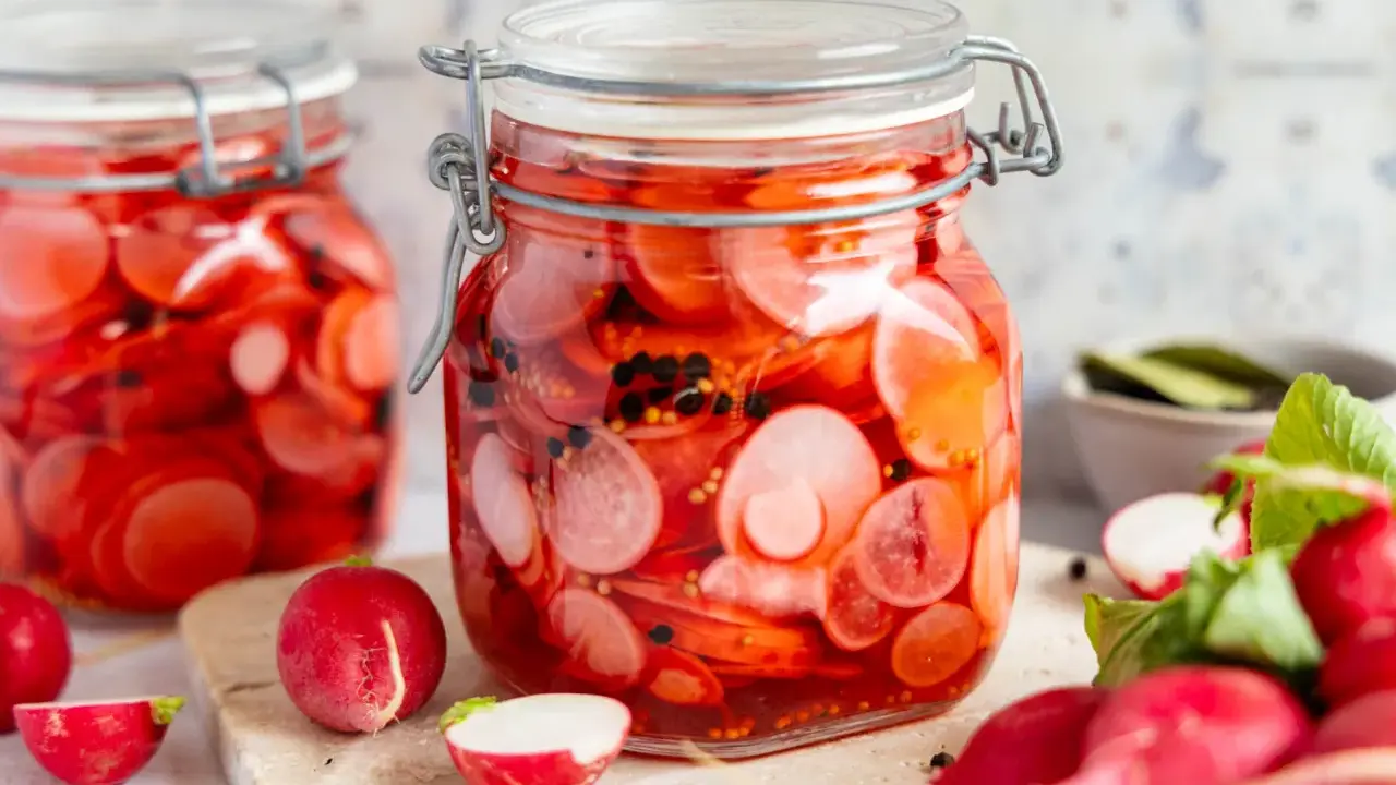 Zwei Gläser mit eingelegten Radieschen, leuchtend rot und mit schwarzen Pfefferkörnern. Frische Radieschen liegen daneben.
