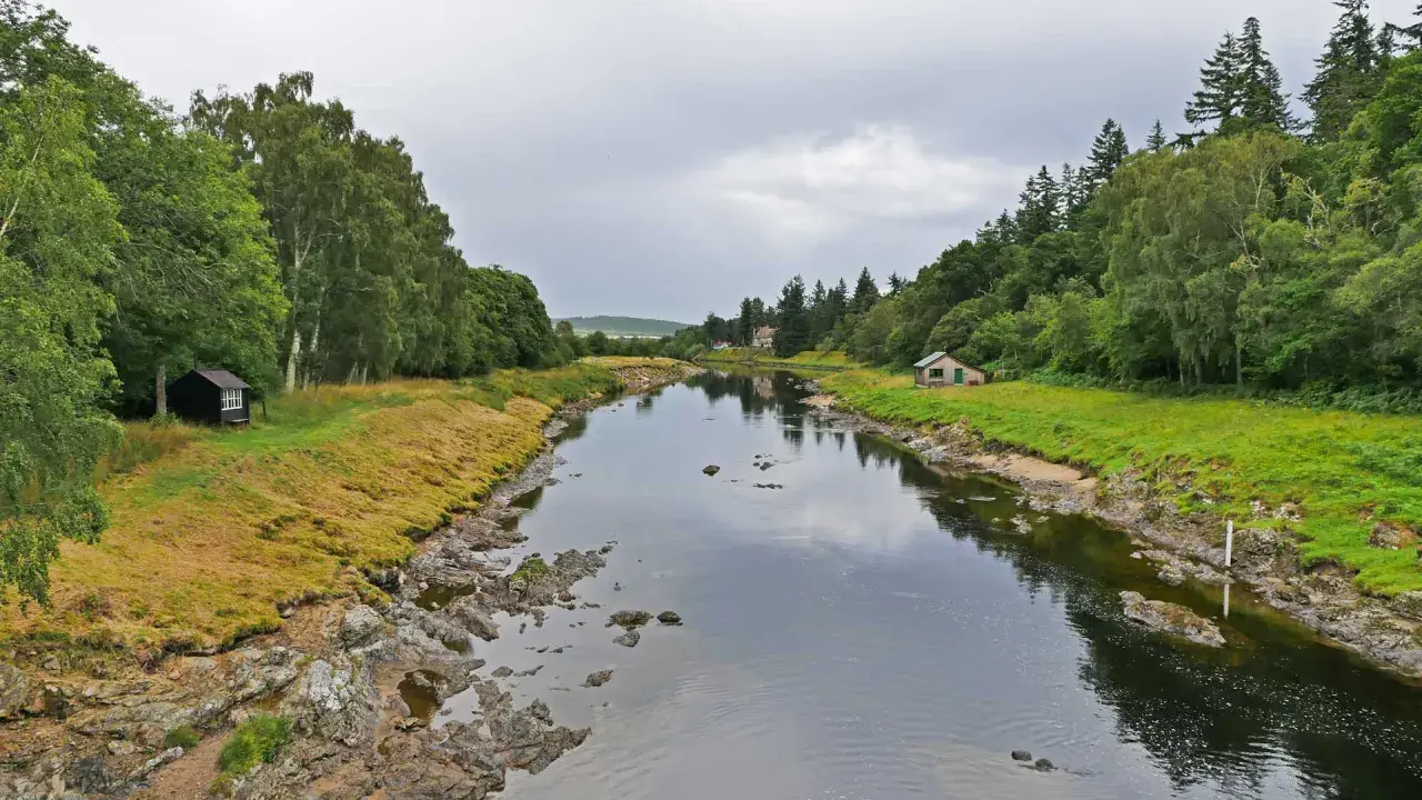 Ein ruhiger Fluss, der durch eine grüne Landschaft fließt. Kleine Häuser säumen das Ufer, vielleicht Orte, an denen man das schottische Uisge Beatha genießt.