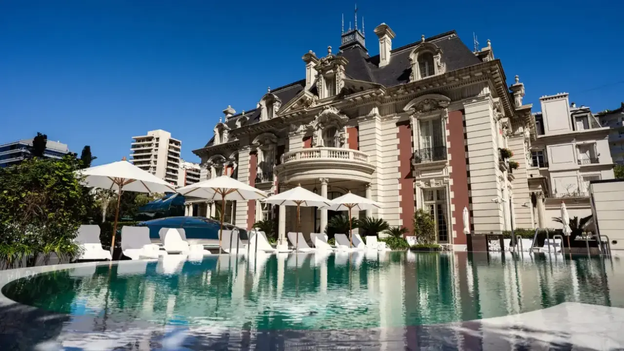 En lyxig pool framför en ståtlig byggnad som påminner om White Lotus Hotel, med solstolar och parasoller.