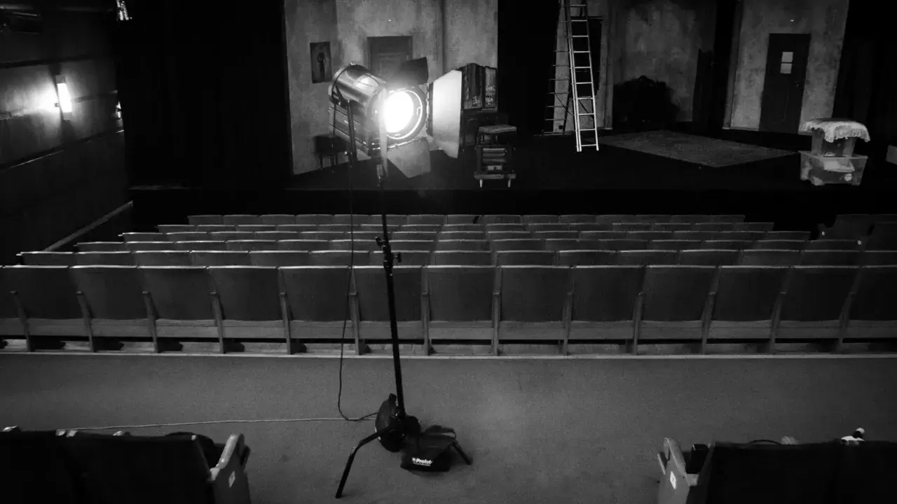 Teatr w czerni i bieli. Na scenie reflektor fresnel oświetla dekoracje, a przed nią puste rzędy siedzeń.