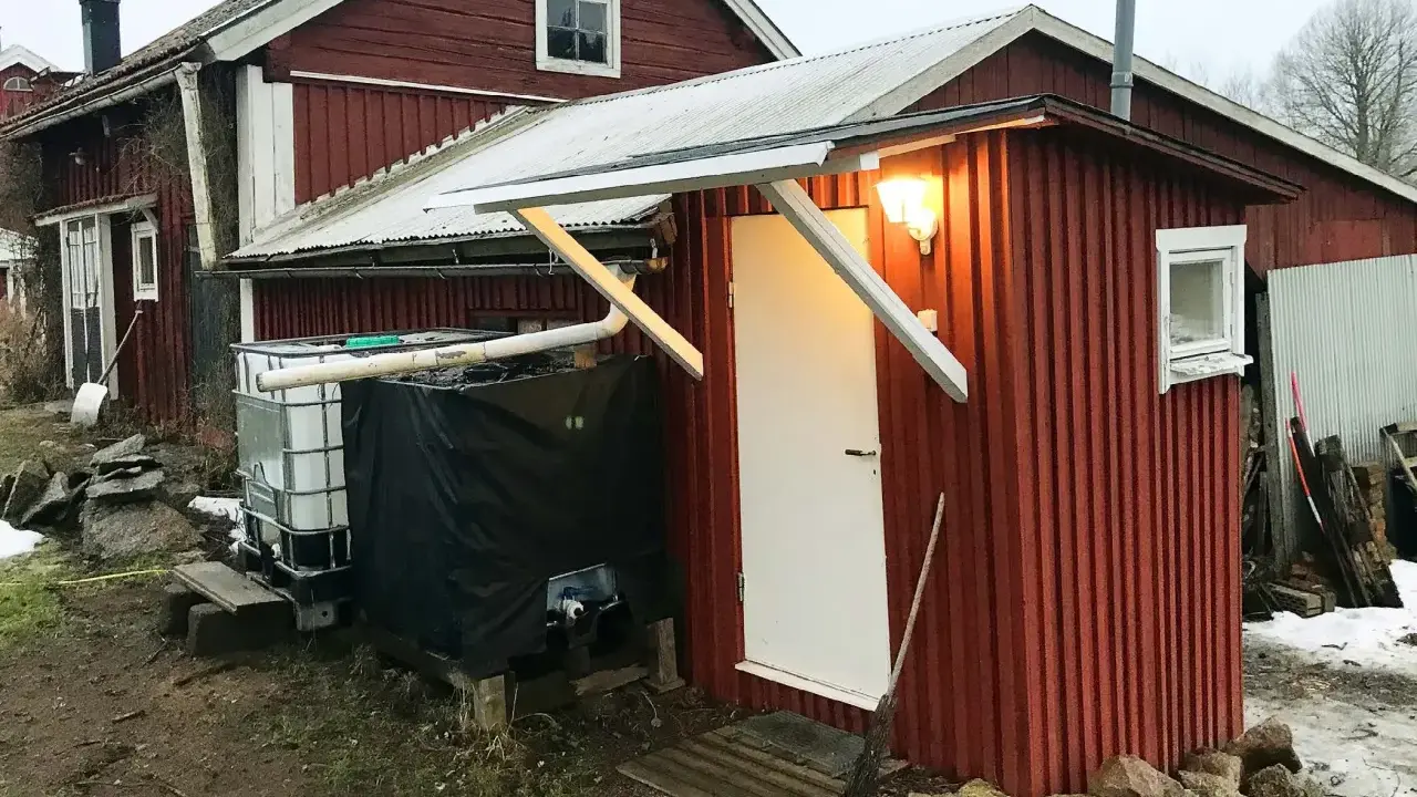 Rött utedass med vit dörr och fönster. Bredvid står en stor tank och en svart presenning. Här följer man utedass regler.
