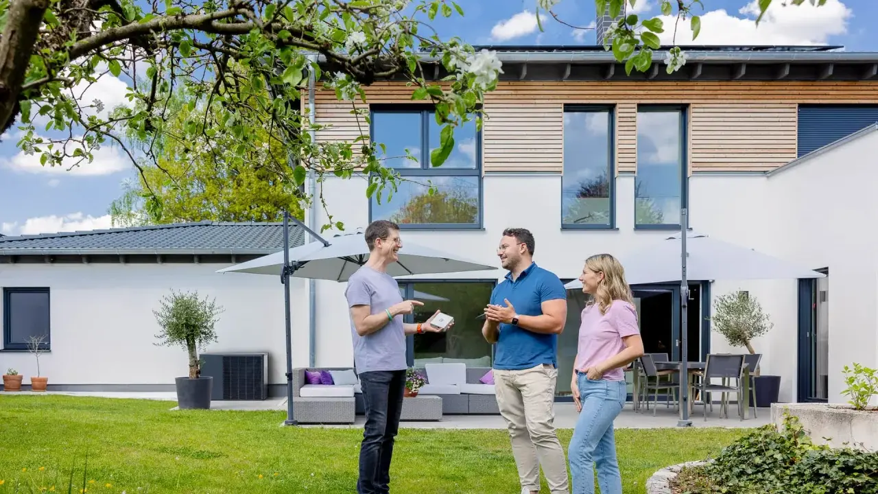 Tre personer diskuterar solcellslösningar på en grönskande trädgård framför ett modernt hus. PFA Solteknik hjälper dig att förverkliga dina drömmar.