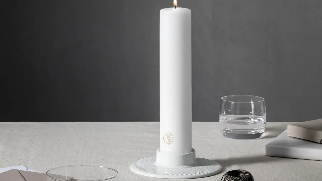 Ett vitt, högt stearinljus med guldig logotyp står i en vit ljusstake. Bredvid finns ett glas vatten, böcker och garn. Olika former av stearinljus kan skapa stämning.