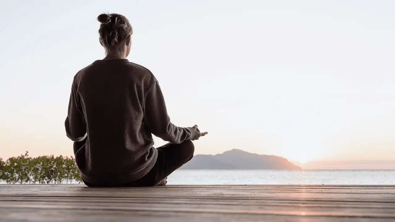 Person sitter i lotusställning på en träbrygga vid havet, med solnedgång i bakgrunden. En stund av mindful yoga.