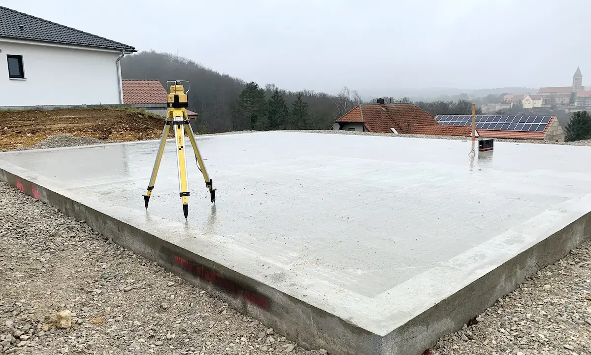 Der **Bodenplatten-Aufbau** ist fast fertig. Ein Nivelliergerät auf einem Stativ steht auf der frisch gegossenen Betonplatte. Im Hintergrund sind Häuser und eine Kirche zu sehen.