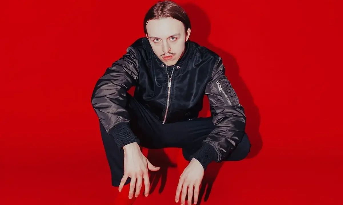 Tommy Cash w czarnej kurtce bomberce i czarnych spodniach kuca na czerwonym tle.
