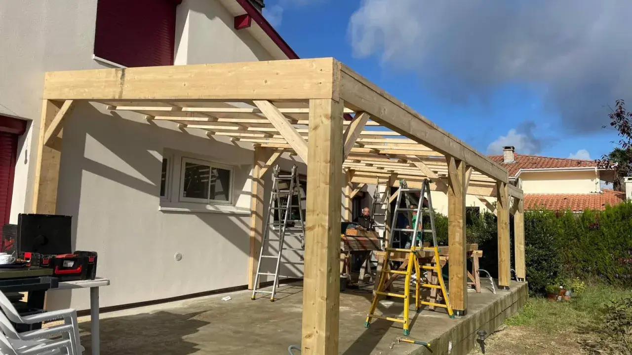Construction d'une charpente sans ferme en bois, avec des poteaux et des poutres, sur une terrasse. Des échelles sont présentes.
