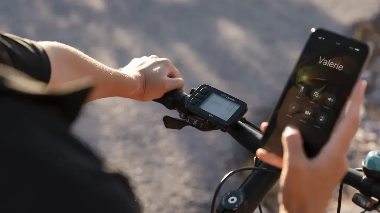 Rowerzysta trzyma telefon, na którym wyświetla się połączenie z Valerie. Obok widoczne są symbole na liczniku rowerowym, pokazujące prędkość i inne dane.