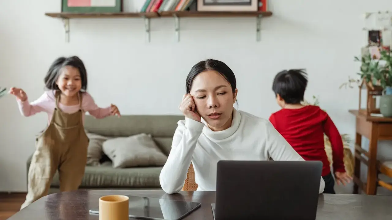 Donna esausta davanti al laptop, mentre i bambini giocano. Un quadro di **parental burnout** in casa.