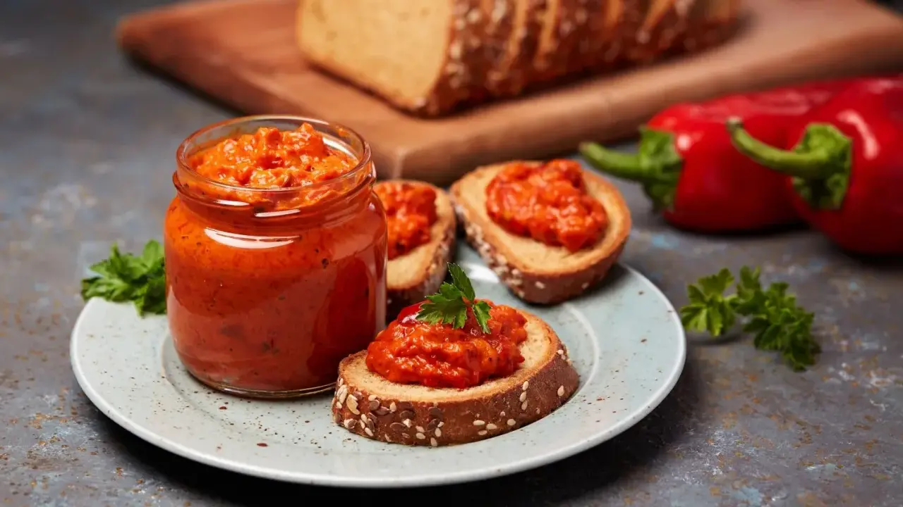 Słoik pełen ajvaru, pysznej pasty z papryki, podany z chlebem i świeżymi paprykami. Ajvar co to? To smak Bałkanów!