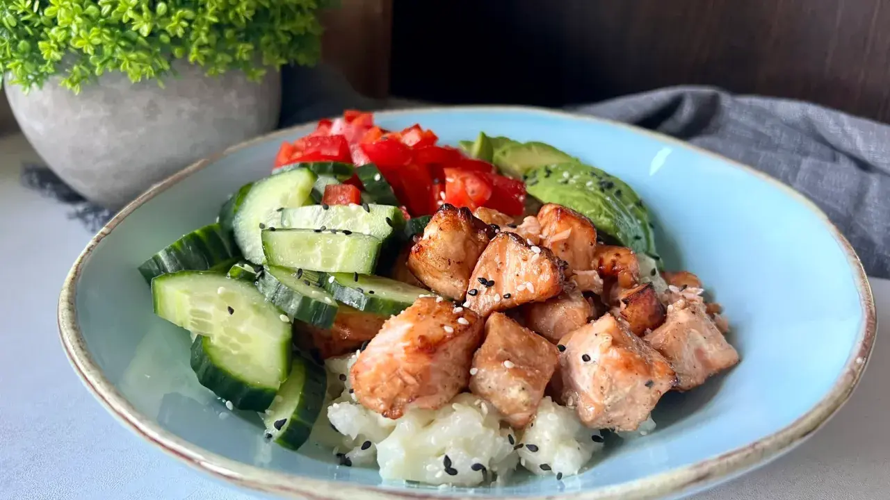 Pyszny bowl z kawałkami pieczonego lososia, świeżym ogórkiem, pomidorem i awokado na ryżu.