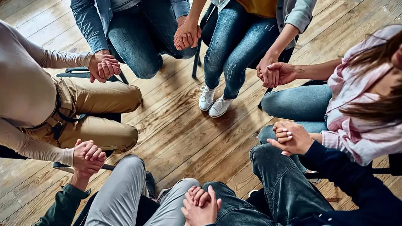 Grupa osób trzyma się za ręce w kręgu, wspierając się nawzajem. Widać tu pracę specjalisty od uzależnień.