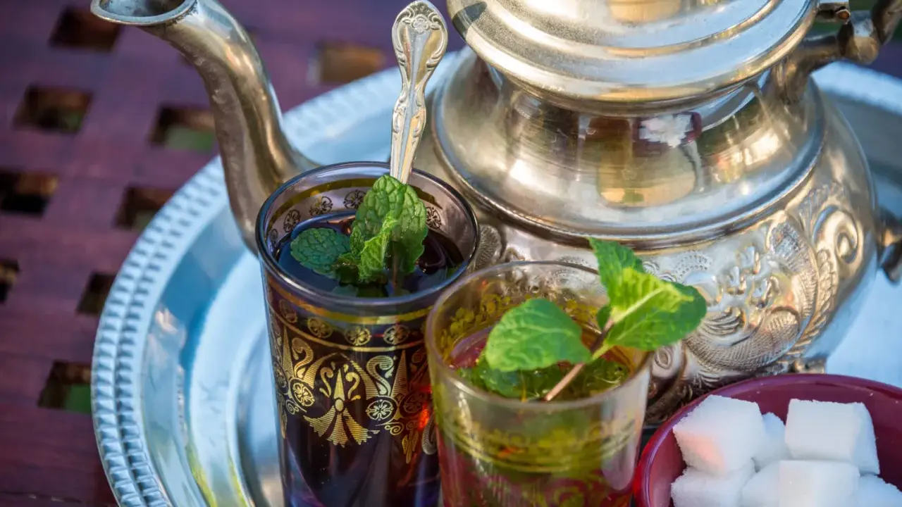 Zwei Gläser mit Minztee, garniert mit frischen Minzblättern, stehen neben einem traditionellen Teekessel und Zuckerwürfeln. Verschiedene Minze Arten verfeinern das Getränk.