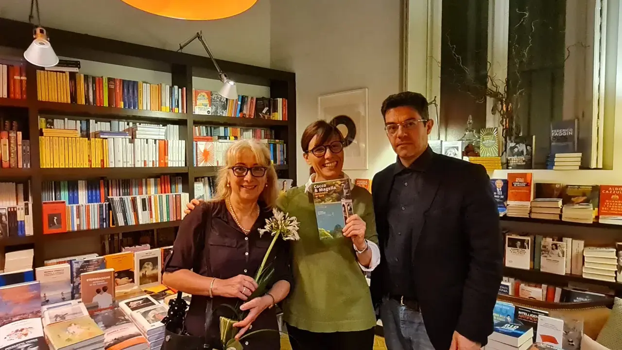 Tre persone sorridono in una libreria, una tiene in mano il libro "Cocco e Magella". Libri in ordine cronologico sugli scaffali.