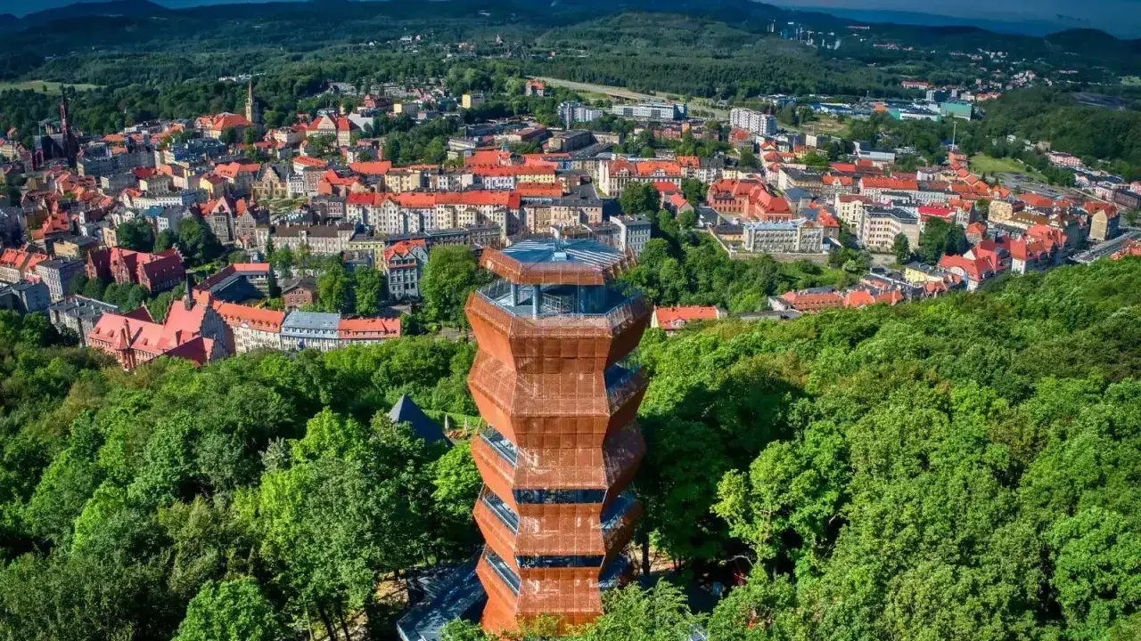 Nowoczesna wieża widokowa w otoczeniu zieleni, z panoramą miasta i Gór Wałbrzyskich w tle.