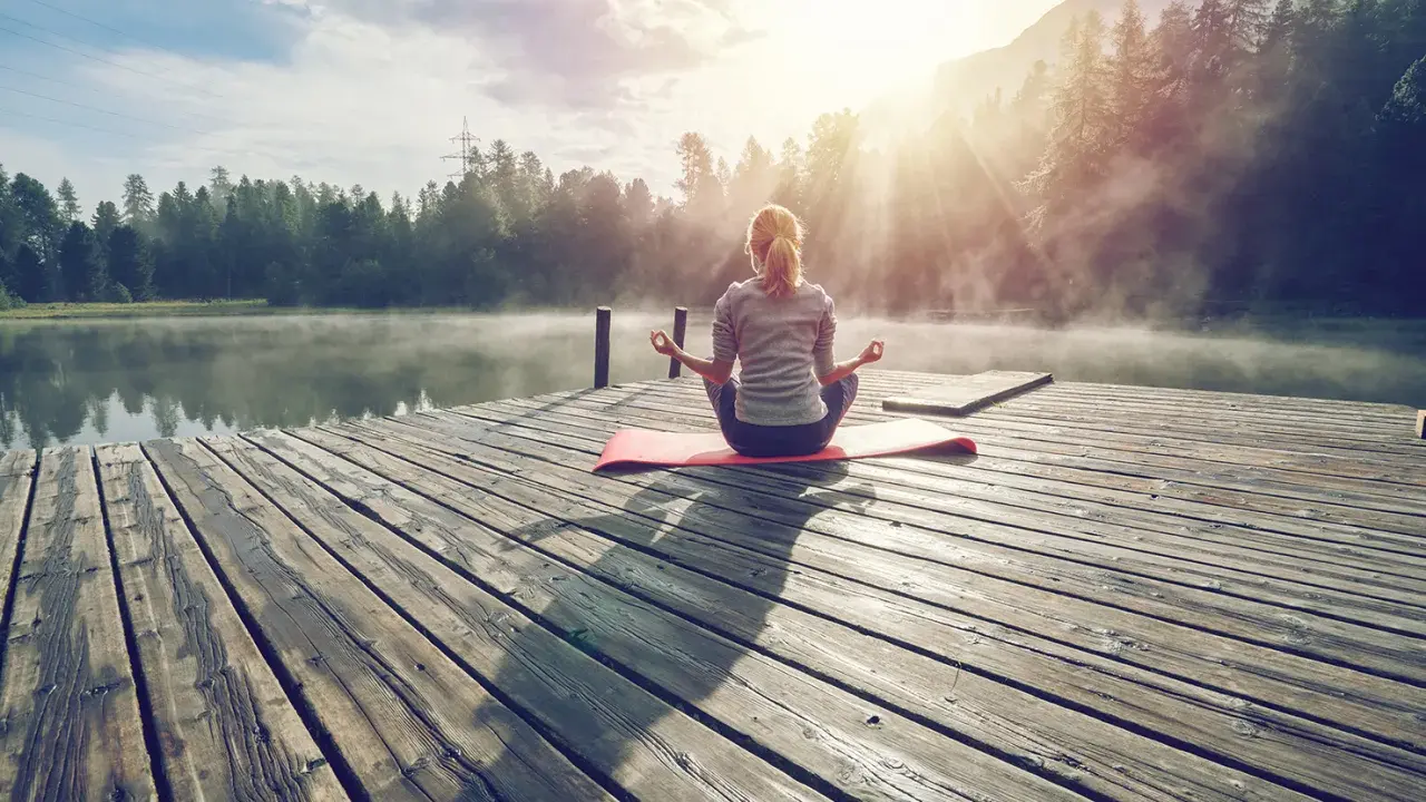 En kvinna med ryggen mot kameran sitter i meditationsställning på en brygga vid en sjö. Solen strålar genom dimman och skapar en magisk stämning. Hon verkar vara helt i nuet.